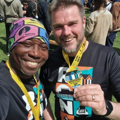 Two men, Tee and Simon, with medals and running race tee shorts, smile after the Worthing Runfest 10K in May 2025